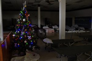 A lit Christmas tree stands in a dark room filled with sleeping cots.