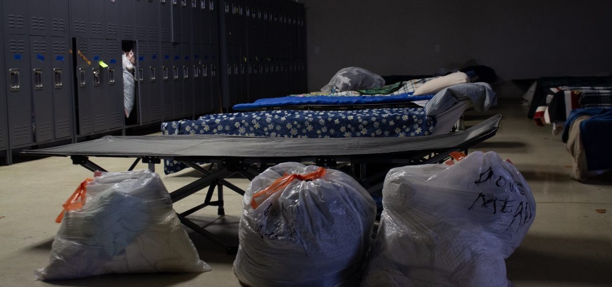 Three plastic bags with clothes inside sit in front of a row of cots. In the background is a set of lockers.