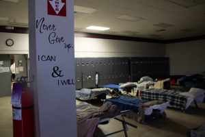 The words "Never Give Up" and "I CAN & I WILL" are printed on a support beam in a room full of cots.