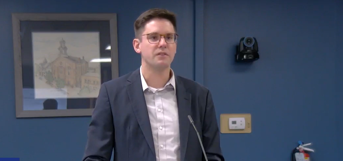 Taylor Koch from Hill Tide Partners gives a presentation on a proposed affordable housing complex in Athens. He's standing at the podium in front of a blue wall.