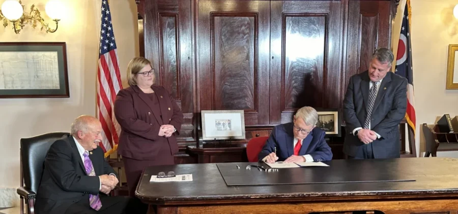 Gov. Mike DeWine signs Senate Bill 109 at a big table with three onlookers