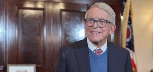 Gov. Mike DeWine smiles while standing in front of a podium microphone.