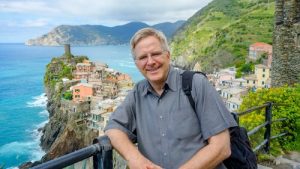 Rick Steves' leaning against railing overlooking a body of water and an Italian town