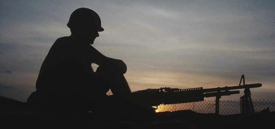 Silhouette of American soldier in Vietnam with high power rifle on ground, chain link fence just beyond him