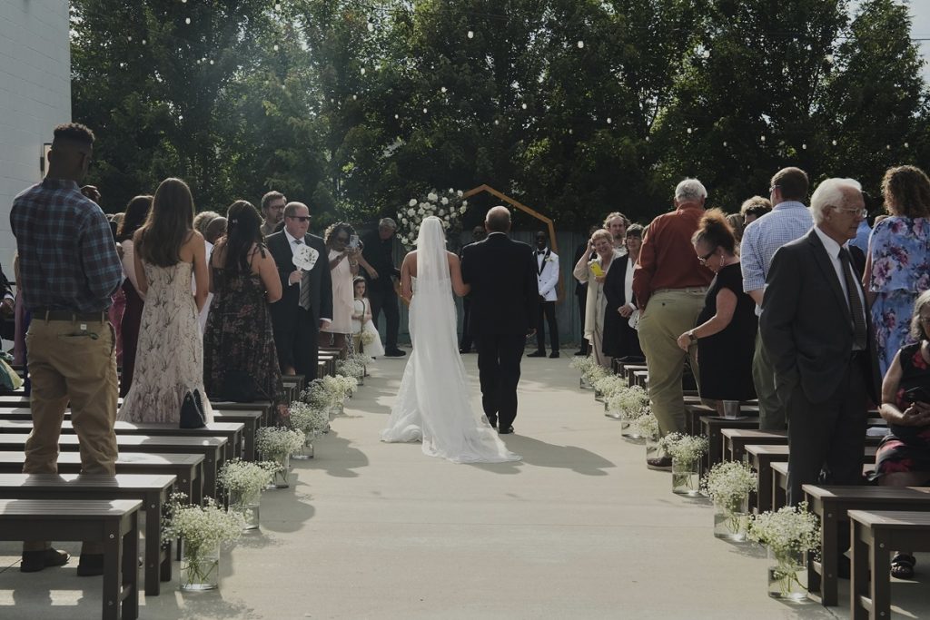 Kayla McDonald walks down the aisle with her father, Chris, during her wedding. Guests hold paper fans to stay cool.