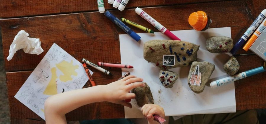A child is seen messing with rocks and art supplies on a wooden table.