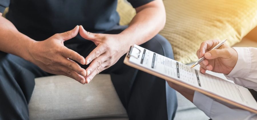 A man sits with his hands together while another person writes on a for clipped to a clipboard.