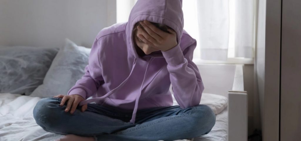 A child wearing a hoodie over their head sits on a bed with their head in their hand.