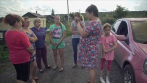 A group of white women and a young girl gather on a dirt road in a residential area. A woman in a floral-patterned blue and pink dress holds a small camera in one of her hands as she speaks to the gathered crowd. A pink car is visible to their right.
