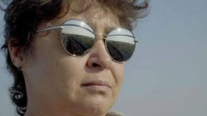 A close-up of a white woman's face wearing round, metallic sunglasses. The lenses reflect railroad tracks stretching into the distance against a bright sky. Her dark hair frames her face.
