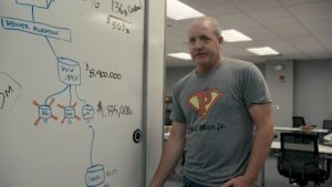 A white man wearing a grey t-shirt with a superhero-style "P" logo stands in front of a whiteboard covered in diagrams and numbers