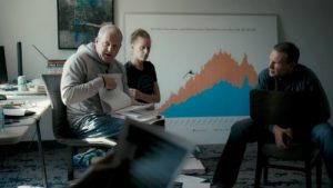 Two men and a woman in an office examine data charts. One man in a grey hoodie speaks passionately while looking through a large stack of papers on his lap.