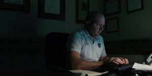 Paul Farrell Jr, works at his computer wearing a white striped polo shirt, in a dimly lit office, focusing on work in front of him.