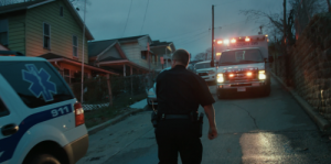 An evening scene shows a police officer walking between an police car and ambulance with flashing lights on a residential street.