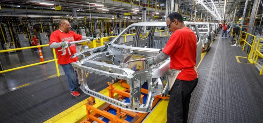 Factory workers stand on the line as the frame of a vehicle in production comes by.