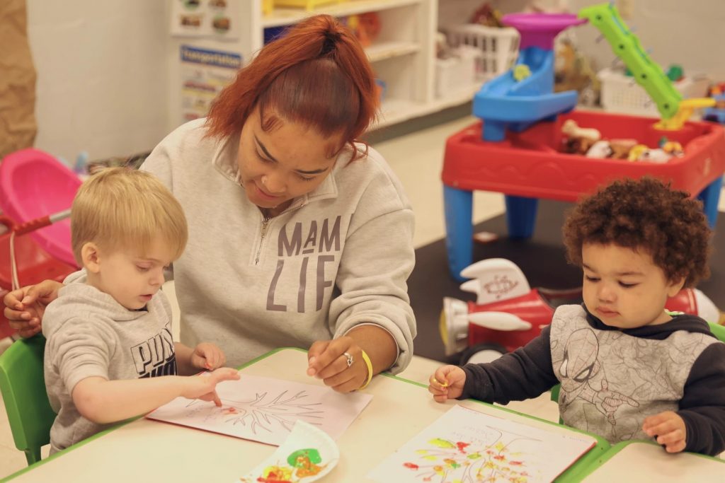 An adult works with two very young kids at a table as part of child care.