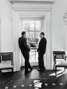 Henry Kissinger and President Richard Nixon stand looking at each other leaning against opposite sides of a doorway.