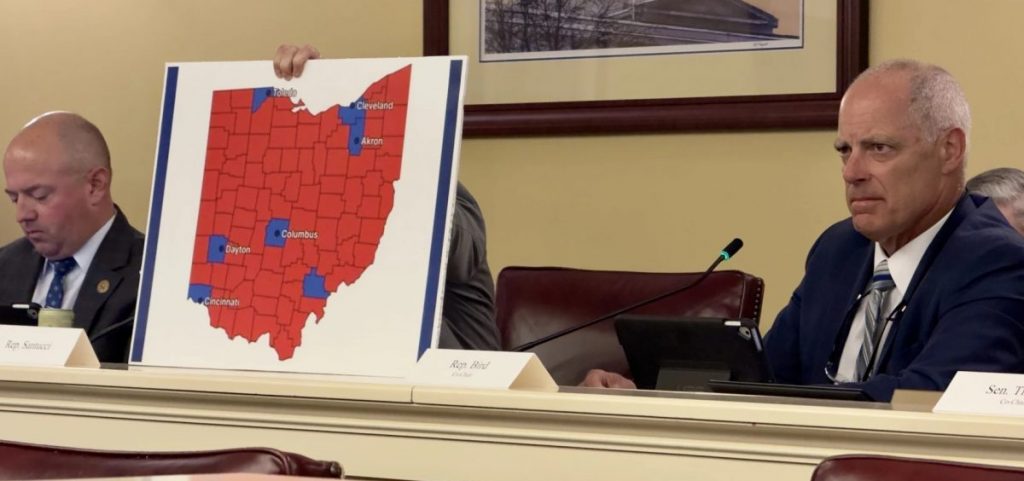 Reps. Brian Stewart, left, and Adam Bird, right, sit in the Joint Committee on Congressional Redistricting, as Nick Santucci holds a map of counties President Trump won in 2024.