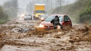 Four cars and a box truck are stranded on a road during a flash flood. A man clings to the hood of his car.