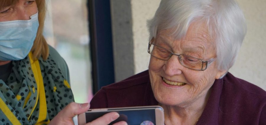A woman wearing a mask holds a phone up to an elderly woman who smiles.