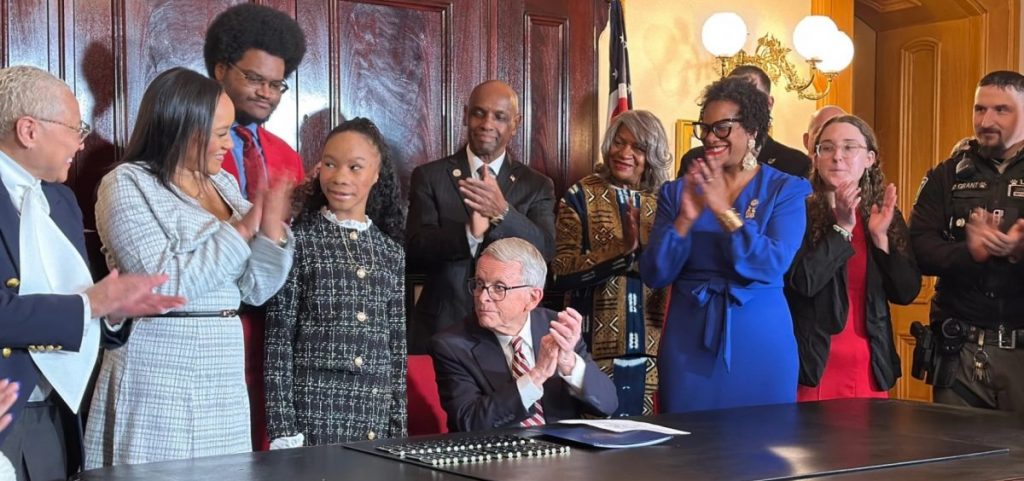 Avery Russell (center) smiles at applause from family and lawmakers gathered to watch Gov. Mike DeWine sign the law that bears her name.