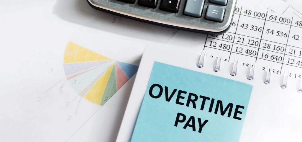 A person is going over finances with a calculator and spreadsheets and a circle graph. There is a post-it note that reads Overtime Pay.
