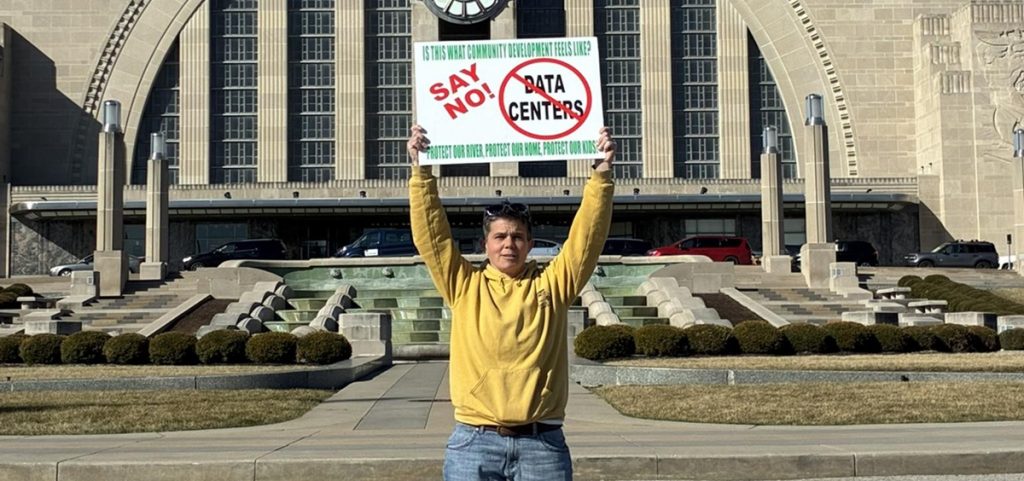 Nikki Gerber, a member of Adams County for Responsible Development, stands with a sign protesting data centers in front of a museum.