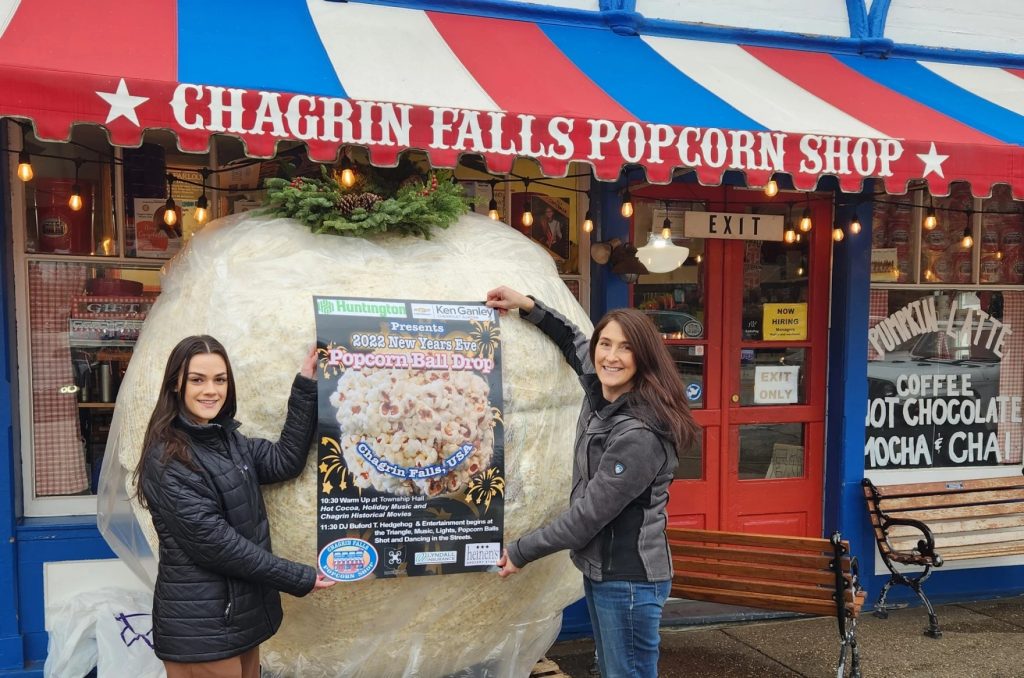 Two women hold a poster for the Popcorn Ball Drop in front of a giant ball of popcorn wrapped in a clear plastic bag.