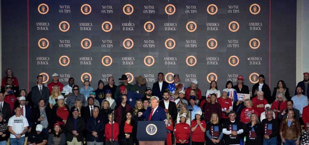 President Donald Trump speak at a podium with a crowd and a backdrop that reads "no tax on tips" behind him.