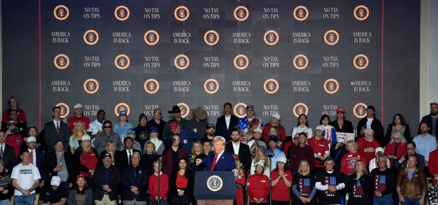 President Donald Trump speak at a podium with a crowd and a backdrop that reads "no tax on tips" behind him.