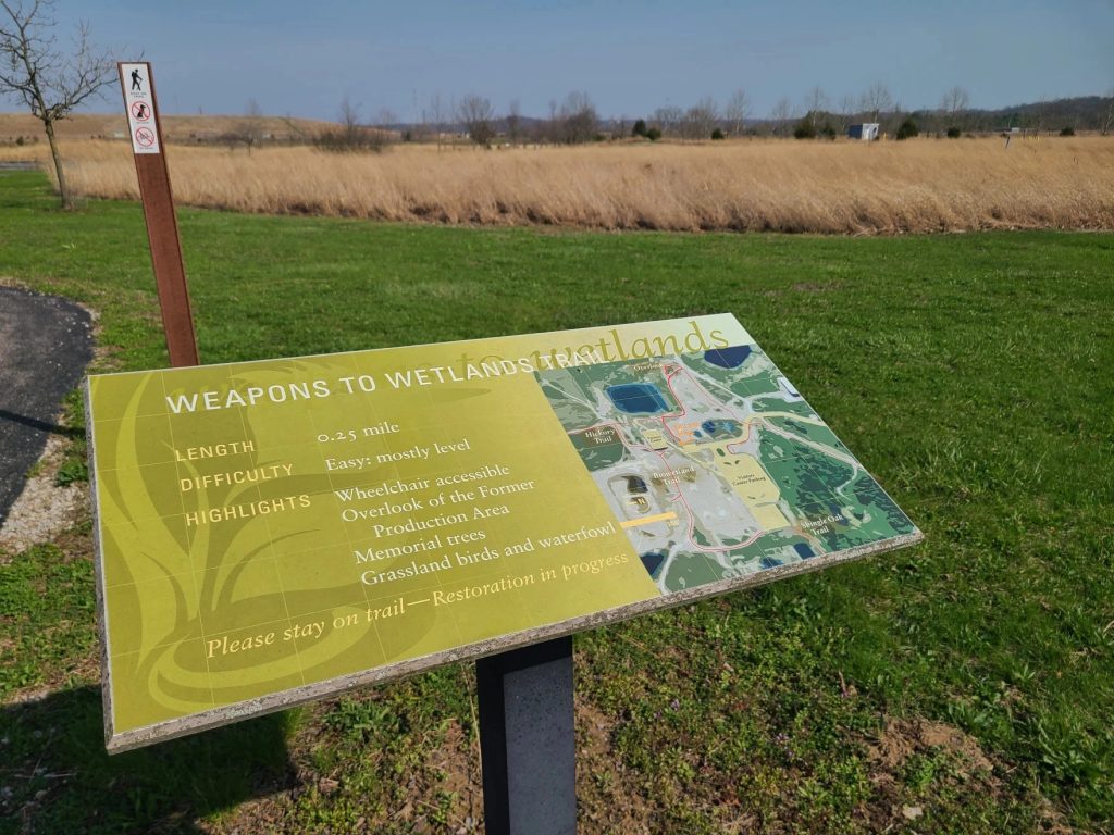 An interpretive sign outlines Fernald's "Weapons to Wetlands Trail." In the upper left corner, the edge of the on-site disposal mound can be seen. It contains around 3 cubic yards of low-grade contaminated waste.