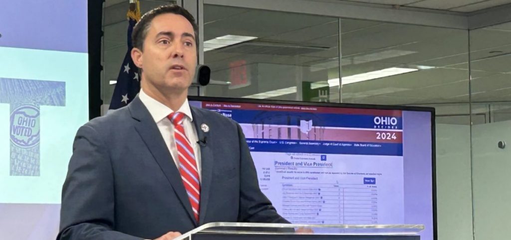 Ohio Secretary of State Frank LaRose speaks at a podium with a screen with data behind him.