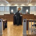 A man stands facing away from the camera and toward three judges sitting in the front of a courtroom.
