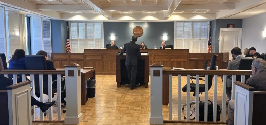 A man stands facing away from the camera and toward three judges sitting in the front of a courtroom.