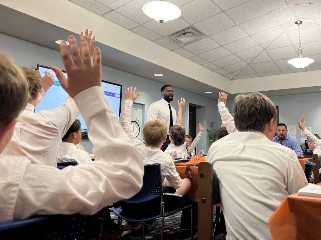 A room full of school boys in white button up shirts and ties sit in a classroom.