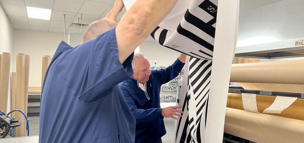 Two men look over a sheet of pressed fabric they are holding in the air together.