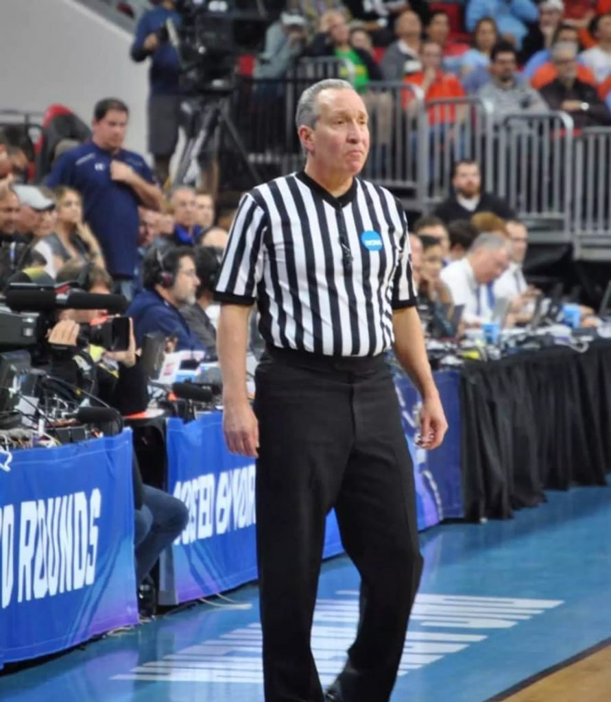 A man stands on a basketball court wearing a striped referee uniform and black pants.