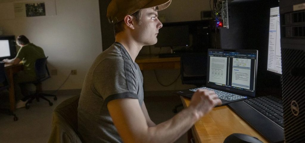 A young man sits at a desk with a computer and a laptop in front of him. He is wearing a hat.