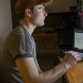 A young man sits at a desk with a computer and a laptop in front of him. He is wearing a hat.