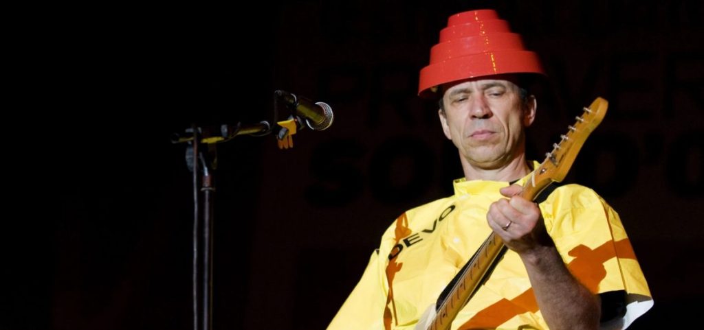 Devo guitarist Bob Mothersbaugh performs on stage wearing a yellow outfit and a red flowerpot looking thing on his head.