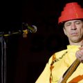 Devo guitarist Bob Mothersbaugh performs on stage wearing a yellow outfit and a red flowerpot looking thing on his head.