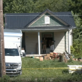 Two trucks are parked outside of an older looking house with junk being piled in front. Two people look through the front window while wearing gloves.