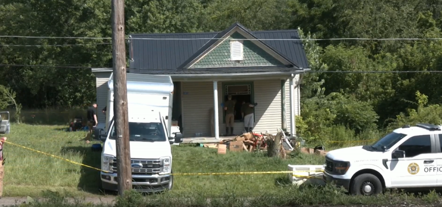 Two trucks are parked outside of an older looking house with junk being piled in front. Two people look through the front window while wearing gloves.