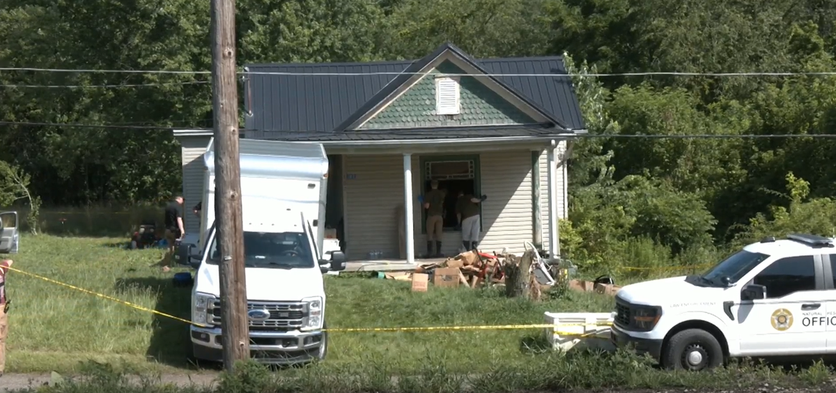 Two trucks are parked outside of an older looking house with junk being piled in front. Two people look through the front window while wearing gloves.