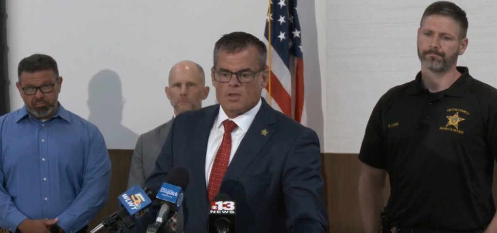Ohio Attorney General Andy Wilson speaks at a podium to the press with two men standing to the left and one standing to the right.