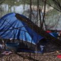 A tent sits along the river in a wooded area.