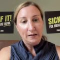 A woman talks on a video chat with two signs behind her reading Sick of It Fix Health Care Now protectourcare.org
