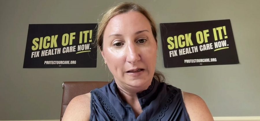 A woman talks on a video chat with two signs behind her reading Sick of It Fix Health Care Now protectourcare.org