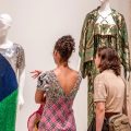 Two people look at three dresses on display at a museum.