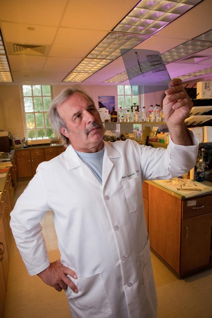John Kopchick, wearing a white lab coat, holds up an xray image in a lab to look at.